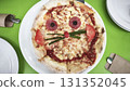 Creative kids pizza in the shape of a cat with eyes made of salami sausage, olives, onions, and tomatoes, topped with melted cheese on a white plate, top view. Kids food.  131352045