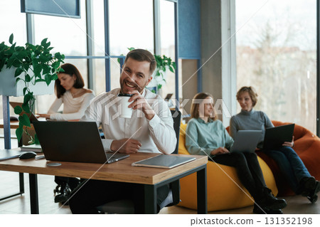 Man is smiling, sitting by the table with laptop. People are working in the office with bean bags chairs in it 131352198