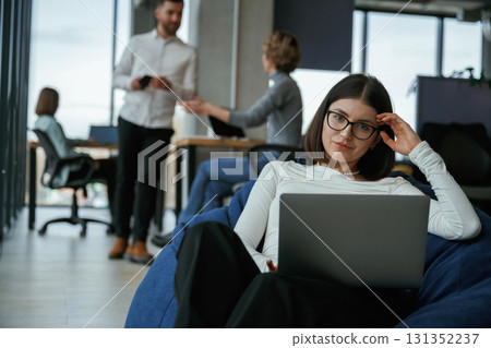 Woman in glasses is sitting with laptop. People are working in the office with bean bags chairs in it 131352237
