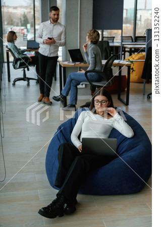 Smart woman is sitting and uses laptop. People are working in the office with bean bags chairs in it 131352240