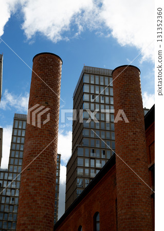 Blend of old and new architecture, two towering brick chimneys juxtaposed with modern high rise buildings. Contrast highlights transformation of urban landscapes, combining historical elements Blend of old and new architecture, two towering brick chimneys juxtaposed with modern high rise buildings. Contrast highlights transformation of urban landscapes, combining historical elements 131352360