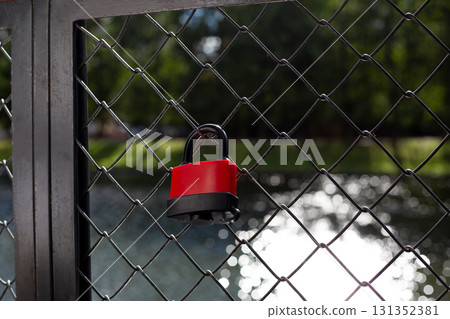 Close up shot of red padlock attached to metal fence with blurred background. The shiny surface of the water and trees provide calm, nature filled atmosphere behind the security feature Close up shot of red padlock attached to metal fence with blurred background. The shiny surface of the water and trees provide calm, nature filled atmosphere behind the security feature 131352381