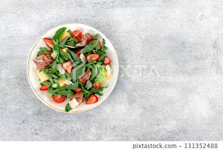 Italian arugula salad with prosciutto, strawberries, melon, cheese on gray background. Top view. Copy space. Italian arugula salad with prosciutto, strawberries, melon, cheese on gray background. Top view. Copy space. 131352488