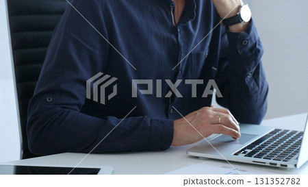 Businessman wearing a blue shirt is typing on a laptop with financial reports and a tablet on his desk, working diligently in a modern office environment. Business people concept Businessman wearing a blue shirt is typing on a laptop with financial reports and a tablet on his desk, working diligently in a modern office environment. Business people concept 131352782