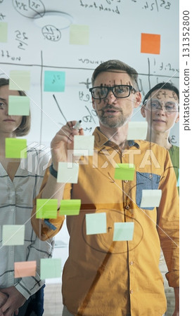 Professional team brainstorming, sharing strategy on glass whiteboard with colorful sticky notes, working collaboratively in bright office space 131352800