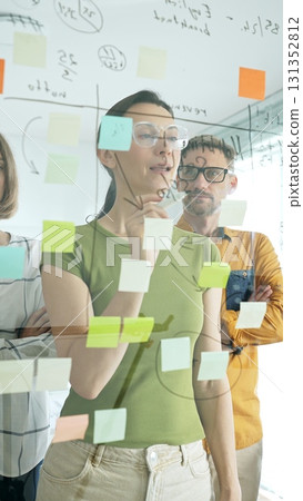 Professional businesswoman wearing glasses writing on glass board, marking strategic plan with sticky notes while colleagues brainstorming in background 131352812