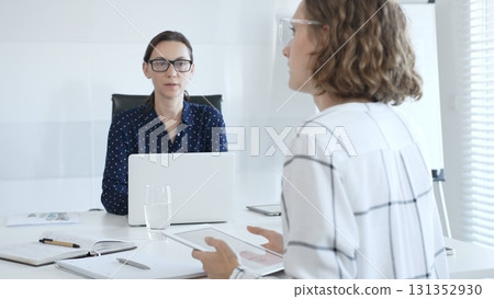 Professional women collaborating at white workspace, reviewing digital content on tablet and laptop during strategic work session 131352930