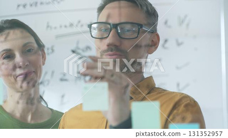 Professional team members brainstorming strategy, writing notes on transparent whiteboard with colorful sticky papers during collaborative meeting. Business people concept 131352957