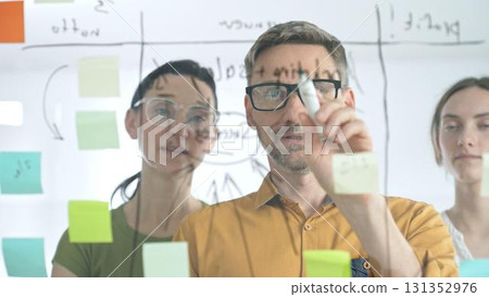 Business colleagues brainstorming, writing strategy notes on glass board, collaborating with colorful sticky notes in creative workplace meeting. Teamwork concept Business colleagues brainstorming, writing strategy notes on glass board, collaborating with colorful sticky notes in creative workplace meeting. Teamwork concept 131352976