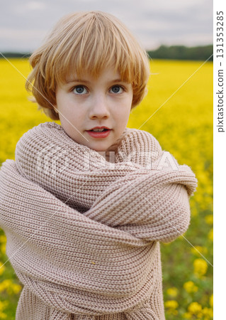 Smiling girl in oversized beige sweater hugging herself in yellow flower field 131353285