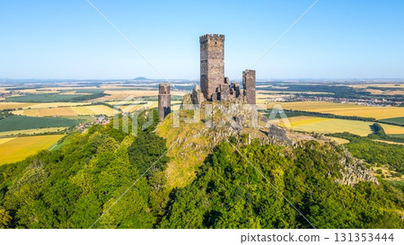 Perched high in the Central Bohemian Uplands, Hazmburk Castle ruins rise on a rocky hill amid lush greenery and farmland, displaying medieval architecture against a clear blue sky. Perched high in the Central Bohemian Uplands, Hazmburk Castle ruins rise on a rocky hill amid lush greenery and farmland, displaying medieval architecture against a clear blue sky. 131353444