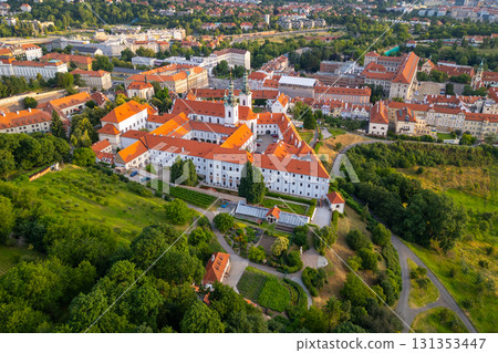 Aerial view of Strahov Monastery bathed in warm sunrise light, surrounded by lush greenery and the cityscape of Prague during the early morning hours. A stunning sight showcasing architectural beauty. 131353447