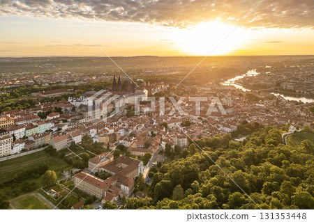 Golden sunlight illuminates Prague Castle, surrounded by historic buildings and lush greenery. The vibrant colors of sunrise create a breathtaking backdrop for this iconic skyline. 131353448