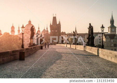 Charles Bridge in Prague glows in the soft light of sunrise. Visitors stroll along the cobblestone path, surrounded by historic statues and breathtaking architecture. 131353450