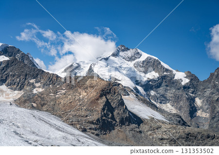 Piz Bernina rises prominently in the Swiss Alps, showcasing its snow-capped peak and glacier. The surrounding rocky landscape highlights the beauty of this alpine region on a clear day. 131353502