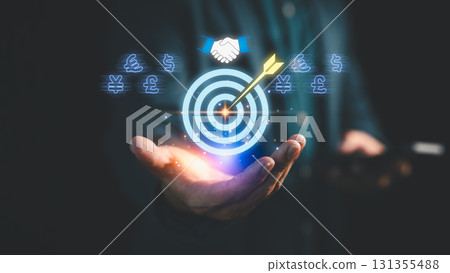 Businessman holding digital target with arrow, currency symbols, and handshake icon, representing financial goal achievement, online business, investment success and global business strategy. 131355488