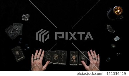 Human hands with rings are positioned over a dark tarot card spread, surrounded by crystals, small spheres, and a lit candle on a black surface, viewed from above. 131356429
