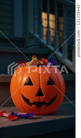 A classic orange plastic jack-o'-lantern bucket filled with colorful wrapped candies sits on a dark wooden porch step at night, with a glowing window in the background. 131356442