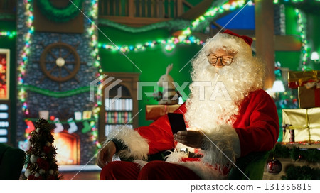 Upbeat man dressed as Santa Claus looks at letters received from kids on smartphone screen. Happy person in Santa costume reads wish lists written by children for Christmas on mobile phone, camera A 131356815