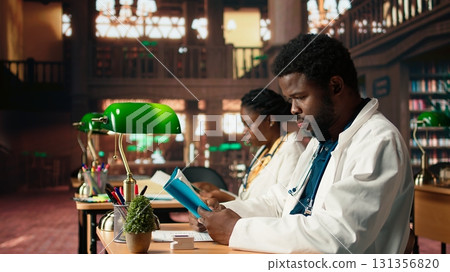 Medical student reading a book and reviewing notes from the library database, focusing on medical research. Young man reflects dedication to healthcare career and academic goals. Camera A. Medical student reading a book and reviewing notes from the library database, focusing on medical research. Young man reflects dedication to healthcare career and academic goals. Camera A. 131356820