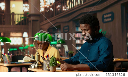 African american pupil underlining key words and phrases at the library, creating a summary with the highlighter pen at a university campus study space. Academic research for exams. Camera B. 131356826