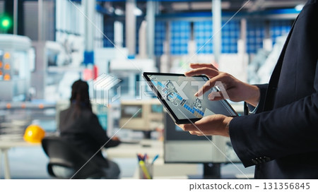 Photovoltaics factory technician monitoring system performance on tablet. Solar panels manufacturing plant worker using device to track energy production and efficiency, camera A close up 131356845