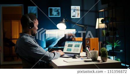 Financial manager doing remote accounting work with music on headset, solving his tasks on laptop at home. African american guy multitasking with his favorite playlist to help him focus. Camera A. 131356872