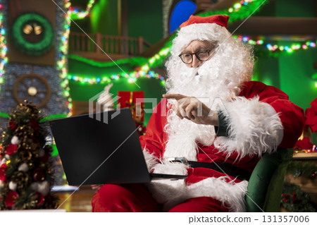 Santa using laptop in festive decorated North Pole workshop to check heartfelt messages from kids worldwide. Senior man wearing Xmas costume reviews wishes sent by children via email using notebook 131357006
