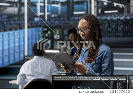 Data center admin using tablet to monitor system performance, identifying bottlenecks. Woman in server room using device to troubleshoot errors, implementing optimizations 131357050