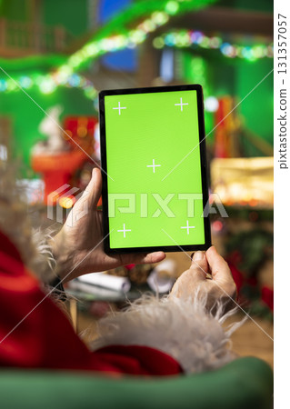 Close up of Father Christmas using mockup tablet to monitor toy shipments during winter holiday season. Man playing Santa Claus character follows gift deliveries with green screen device 131357057