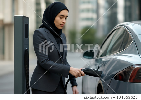 AI generated image of a Middle Eastern businesswoman wearing a stylish pantsuit while charging her electric car at a public station in a modern city center. Sleek, professional, and urban lifestyle. 131359625