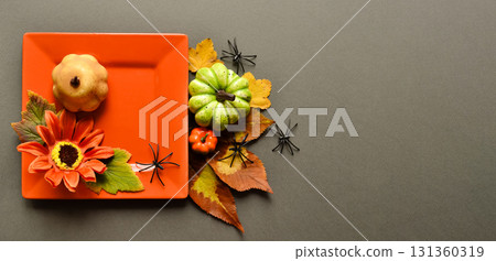 An orange square plate with a sunflower and pumpkin on a gray background with pumpkins, leaves, and spiders. Autumn composition. 131360319