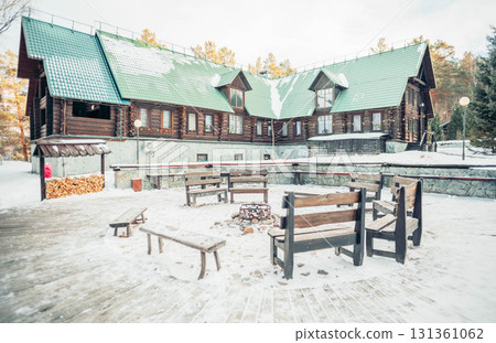 A cozy wooden lodge in the countryside surrounded by snow-covered mountains. A fire pit with benches is in the foreground, creating a peaceful vacation atmosphere. 131361062