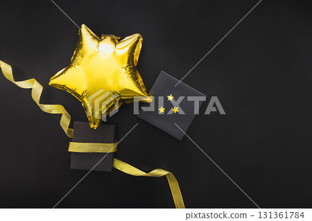 A golden star-shaped balloon rests beside a black gift box with gold stars. A gold ribbon curls around the items on a black background, symbolizing Black Friday sales. 131361784