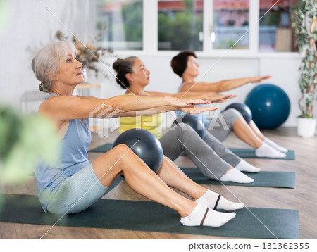 Old woman practicing pilates sitting with softball between knees in light fitness room 131362355