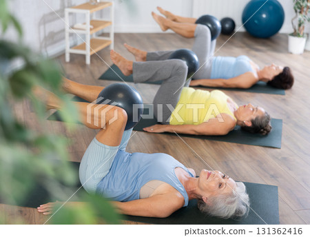 Old woman practicing pilates with softball between legs in light fitness room Old woman practicing pilates with softball between legs in light fitness room 131362416