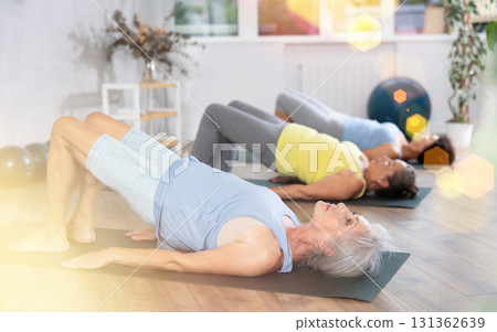 Elderly woman in company of hatha yoga lovers performs bridge pose, setu bandha sarvangasana Elderly woman in company of hatha yoga lovers performs bridge pose, setu bandha sarvangasana 131362639