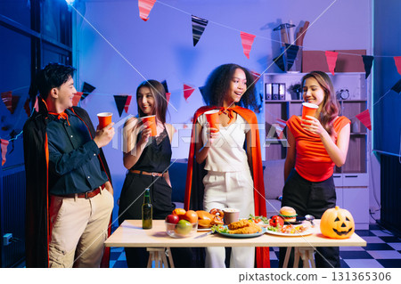 Joyful friends in vibrant Halloween costumes share drinks and laughter at a festive indoor party. Joyful friends in vibrant Halloween costumes share drinks and laughter at a festive indoor party. 131365306
