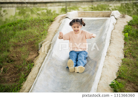 Happy child sliding down a playground slide in a park, enjoying outdoor playtime, smiling and having fun on a sunny day with green surroundings 131366360
