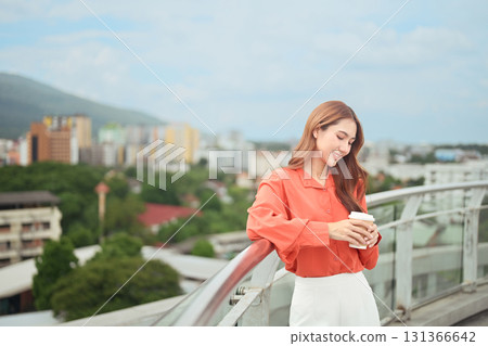 Smiling woman holding a takeaway coffee cup while leaning on a rooftop railing with a blurred cityscape background 131366642
