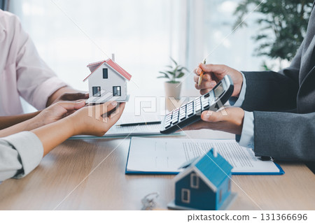 Close up of real estate agent handing house model to client with calculator and contract paperwork on desk, concept of mortgage loan, home ownership, property investment and housing agreement. Close up of real estate agent handing house model to client with calculator and contract paperwork on desk, concept of mortgage loan, home ownership, property investment and housing agreement. 131366696
