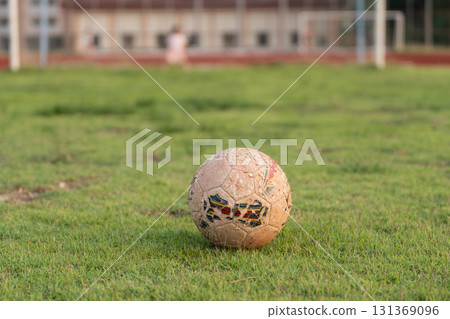Worn Soccer Ball on Grass Field in Soft Evening Light with Blurred Background of Soccer Goal and Children Playing in Distance 131369096
