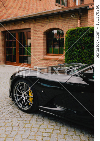 Front side of luxury black sports car with yellow brake calipers, parked on cobblestone driveway in front of brick house with wooden windows and hedge. Sleek black sports car, yellow brake calipers 131370260