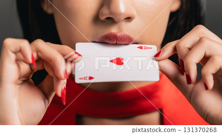 Close-up of Woman with Red Heart Playing Card Between Lips, Wearing Bright Red Outfit, Engaging and Dramatic Mood, Captivating Beauty and Intrigue 131370268