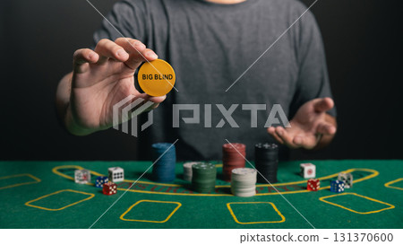 Close-Up of a Person Holding a Casino Chip Marked as Big Blind While Playing Poker at a Green Felt Poker Table with Various Chips and Dice Close-Up of a Person Holding a Casino Chip Marked as Big Blind While Playing Poker at a Green Felt Poker Table with Various Chips and Dice 131370600