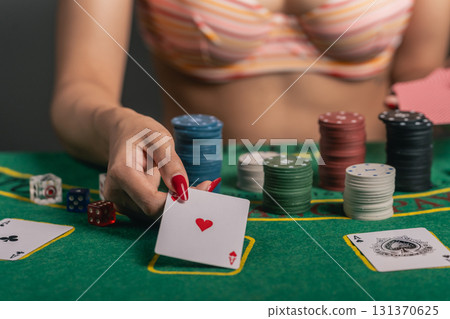Close-up of a Sexy Woman in a Bra Playing Poker, Holding a Playing Card with Heart Symbol on a Green Felt Table Surrounded by Colorful Chips and Dice 131370625