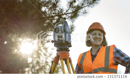 A female geodetic engineer is currently inspecting the construction site, wearing a protective uniform. She is using a high-precision total station to determine the level of the construction, which is A female geodetic engineer is currently inspecting the construction site, wearing a protective uniform. She is using a high-precision total station to determine the level of the construction, which is 131371119
