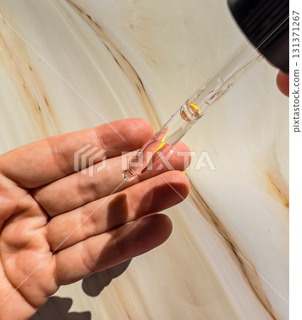 Close-up of a hand applying transparent cosmetic serum with a glass dropper on a bright marble background. Skincare, beauty routine, self-care, and wellness concept. High quality photo 131371267