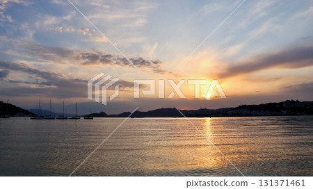 Sun dips below the hills, painting the sky with warm hues as it reflects off the gentle sea, with sailboats silhouetted on the tranquil water. 131371461