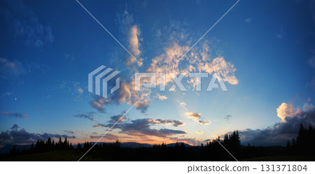 Serene evening sky with hues of blue and soft pink clouds as sun sets. Horizon glows gently, casting light over silhouetted pine trees and rolling hills, creating peaceful and expansive landscape. 131371804